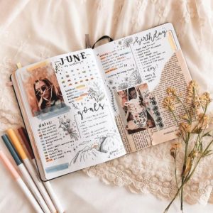  An open elegant vintage themed June bullet journal spread, with illustrations, tasks, and goals on bed with markers and dried flowers